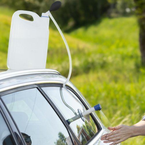 Mycie rąk w plenerze. Widoczny jest kran kempingowy, przymocowany przyssawką do szyby samochodu, korzystając z wody w pojemniku za pomocą zestawu samozasysającego.